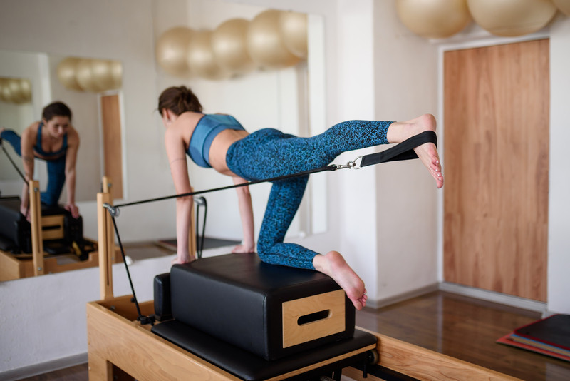 A young woman in sportswear does Pilates on a reformer, lifting her leg up.